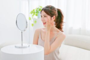 woman with brunette ponytail touches her eye area smiling and looking into mirror
